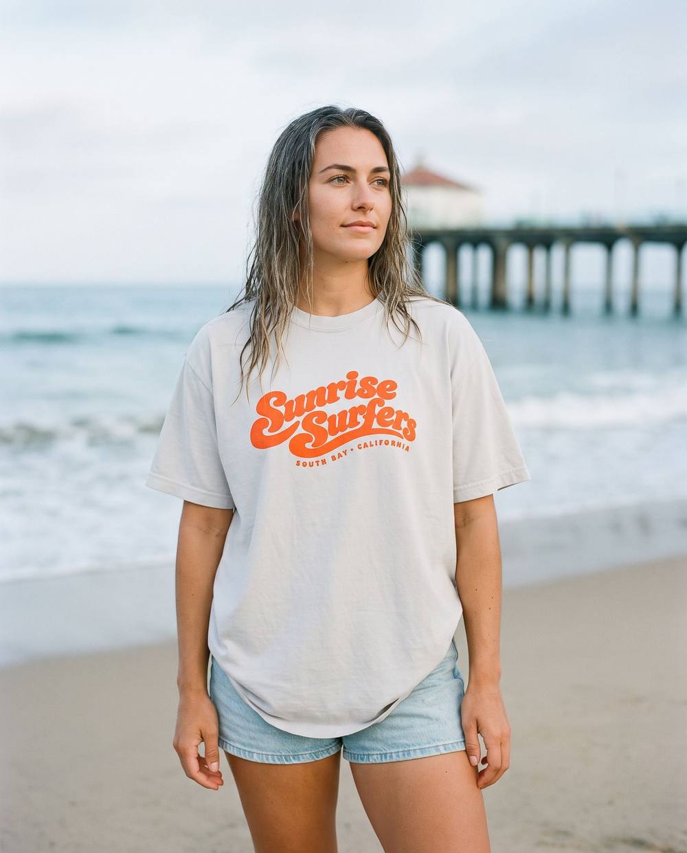 Woman on the beach in the Sunrise Surfers Script Tee, pale grey with orange retro script across the chest.